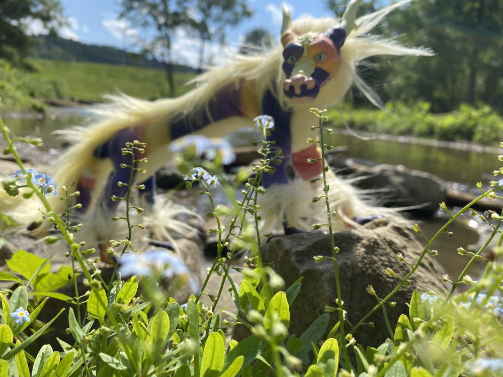 A purple and orange sunrise colored eastern dragon art doll is out of focus in the background looking at some small flowers that are in focus.