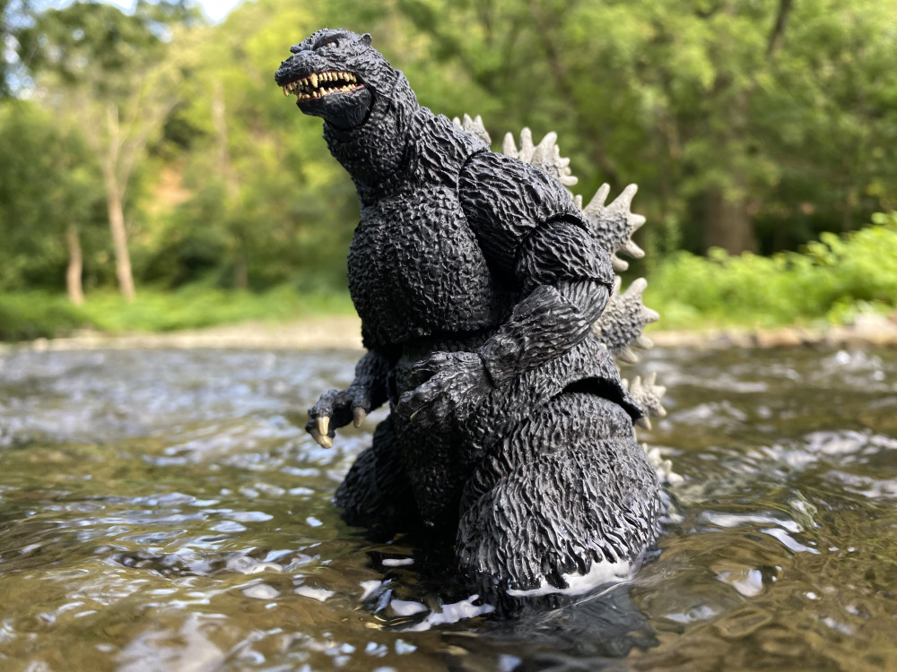 A godzilla toy up to its hips in river water. Green folliage is blurry in the background.