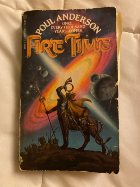 A copy of Fire Time on a white bed. The cover depicts the dark shape of a lion-centaur blowing into a golden horn, the backdrop being a galaxy with various planets around it.