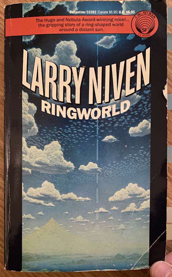 The cover of Ringworld. It depicts a blue sky with swirling clouds and a dotted line far in the distance. The horizon is water with speckles of islands across it, with a pyramid in the far far distance. It say's LARRY NIVEN, RINGWORLD. Along the top is a contrasting red band with the text: The Huge and Nebula Award-winning novel... The gripping story of a ring-shaped world around a distant sun.