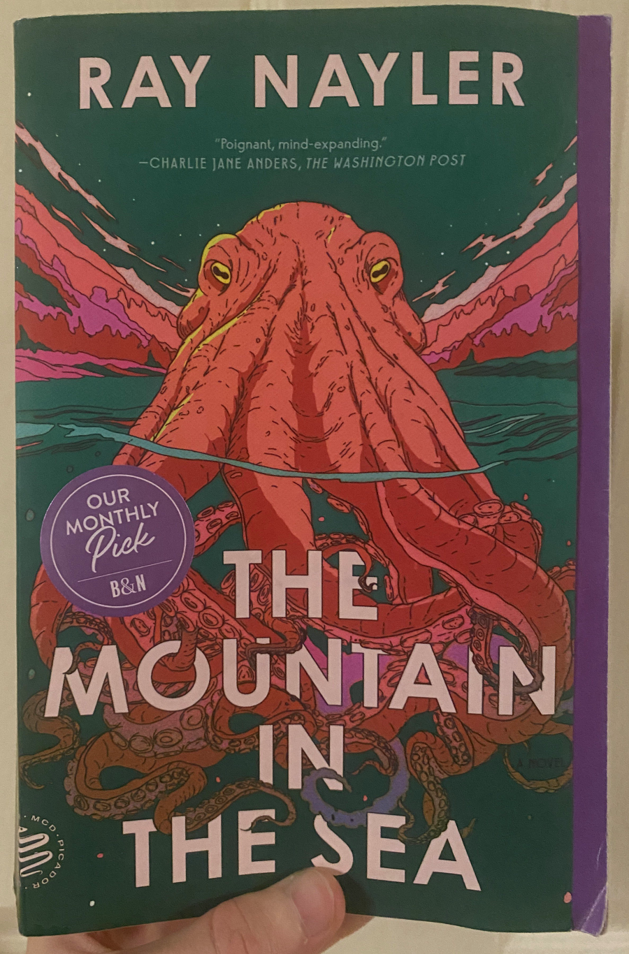 My copy of the book. It features lined unshaded art of an orange octopus looking over the surface of the water. The sky and the water are similar shades of blue-green and the mountains behind the octopus are similar to the color of the octopus. It has a Barnes and Noble monthly pick sticker on it.