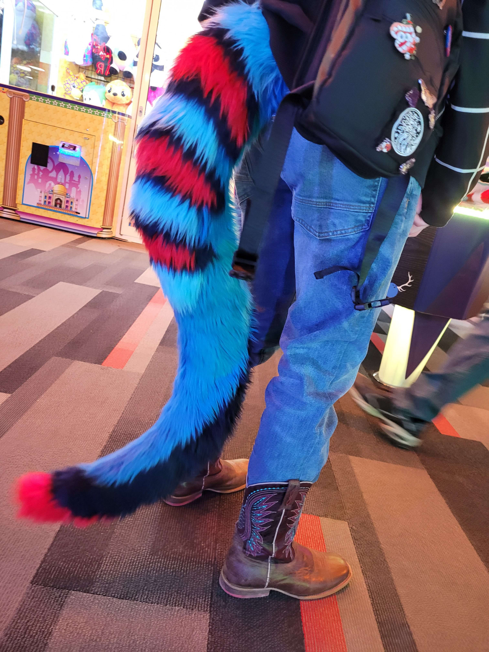 Photograph of a fursuit tail in an arcade setting. The tail has layered blue stripe markings with red details at the top and at the tip.